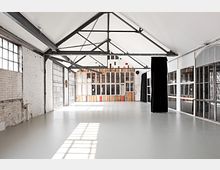 Ein heller, offener Raum mit weiß gestrichenen Wänden und einem schrägen Dach aus sichtbarer Stahlkonstruktion. Die Rückwand besteht aus einer Kombination aus Holzpaneelen und gemischten Fensterelementen, während die seitlichen Wände aus großen Fenstern mit schwarzen Metallrahmen bestehen.