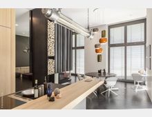 Moderne Wohnküche mit offener Gestaltung, einem Tresen aus hellem Holz, einem Wandregal mit gestapeltem Kaminholz und einer Hängeleuchte aus Metall. Im Hintergrund sind große Fenster mit Jalousien, ein runder Esstisch mit weißen Stühlen und eine gemütliche Sitzecke sichtbar.