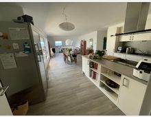 Offene Wohnküche mit modernem Design, bestehend aus einer Küchenzeile mit Arbeitsplatte, Schränken und Küchenutensilien auf der rechten Seite sowie einem Essbereich mit einem langen Tisch und Stühlen im Hintergrund. Der Raum hat große Fenster auf der linken Seite und helle Bodenfliesen, dekoriert mit Blumen und Möbeln.