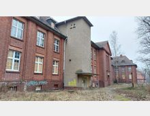 Altes Backsteingebäude mit mehreren Flügeln, großen Sprossenfenstern (teilweise mit Holzplatten verbarrikadiert) und einem zentralen, verputzten Anbau. Die Umgebung ist verwildert, mit kahlen Bäumen und überwuchertem Gras.