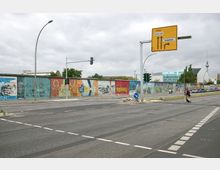 Straßenkreuzung mit Blick auf die bemalte Berliner Mauer (East Side Gallery) und mehrere Verkehrszeichen, darunter ein gelbes Schild mit Richtungshinweisen. Im Hintergrund sind moderne Gebäude, Bäume und der Berliner Fernsehturm zu sehen.