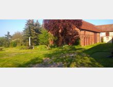 Historisches Backsteingebäude mit roten Dachziegeln, umgeben von einer Wiese und Bäumen, darunter ein Baum mit dunkelrotem Laub; im Vordergrund ein Rasenstück mit vereinzelten Sandflächen und vereinzelten landwirtschaftlichen Gegenständen.