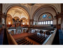 Innenansicht einer Kirche mit weißen Gewölbedecken, roten Backsteinwänden und großen Rundbogenfenstern mit buntem Glas. Zu sehen ist eine Orgel auf einer Empore, hölzerne Kirchenbänke und ein Altarbereich mit dekorativen Elementen.