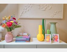 Schlichte Kommode mit Dekoration: Links ein Blumenstrauß in einer rosa Vase, daneben ein Stapel bunter Bücher. Rechts drei dekorative Vasen in Gelb und Grüntönen sowie zwei Bilderrahmen mit farbenfrohen Motiven. Dahinter eine Wand mit Kunstrelief.