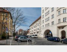 Straßenecke einer Wohngegend mit mehrstöckigen Gebäuden aus beigefarbenem und ockerfarbenem Putz, teils mit Arkaden. Geparkte Autos stehen entlang einer gepflasterten Straße, vereinzelte Bäume ohne Blätter und Verkehrsschilder sind ebenfalls sichtbar.