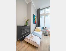 Schmales, modern eingerichtetes Schlafzimmer mit Einzelbett, schwarzer Kommode und hellem Holzboden. Große Fensterfront mit grauen Vorhängen und Heizkörper darunter, daneben ein kleines Beistelltisch-Set und ein farbiges Wandbild.