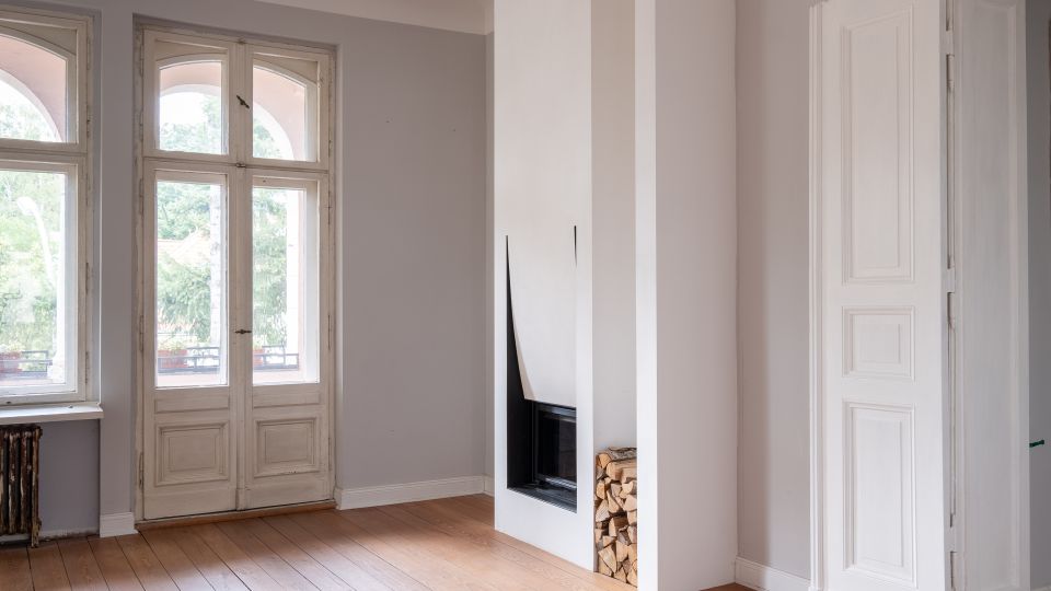 Heller, leerer Wohnraum mit hohen Fenstern und doppelflügeliger Balkontür in weißem Holzrahmen sowie hellem Holzboden. In der Raumecke steht ein moderner, weiß verkleideter Eckkamin mit seitlich gestapeltem Brennholz neben einer hohen weißen Tür.