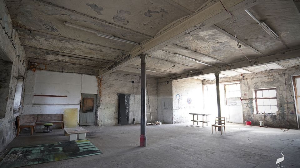 Großer, leerer Industrie- oder Lagerraum mit bröckelnder Betondecke, freiliegenden Trägern und Stützsäulen sowie abgenutzten, weiß gestrichenen Ziegelwänden. An der rechten Seite fallen Fensterlicht und einzelne verstreute Möbelstücke und Gegenstände in dem staubigen Raum auf.