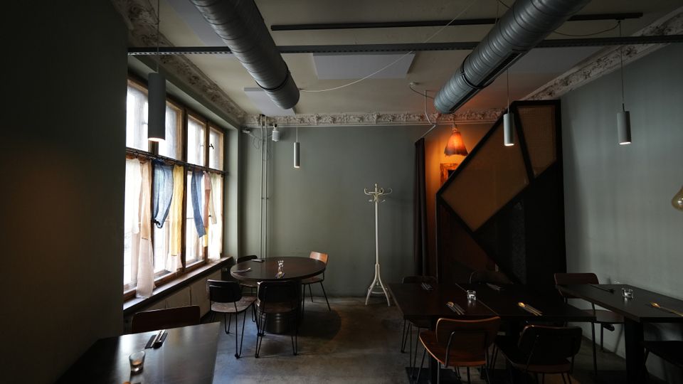 Innenraum eines modernen Restaurants oder Cafés mit grauen Wänden, Betonboden und freiliegenden Lüftungsrohren an der Decke. Links stehen Tische an großen Fenstern mit leichten Vorhängen, rechts weitere Sitzplätze neben einer geometrischen Holzstruktur und hängenden Lampen.