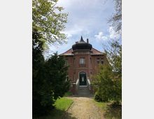 Historisches Backsteingebäude mit einem markanten, kuppelförmigen Turm auf dem Dach, umgeben von Bäumen und Grünflächen. Eine gepflasterte Zuwegung führt zu einer Eingangstreppe mit dunkler Holztür.