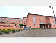 Außenansicht eines rosa Gebäudekomplexes, bestehend aus der „Friedrich-Ebert-Grundschule“ und einem „Stadttheater“. Links ist eine niedrige Mauer mit Pflanzen und Blumenbeeten zu sehen, während rechts ein Eingangsbereich mit mehreren Türen und Fenstern dargestellt ist.