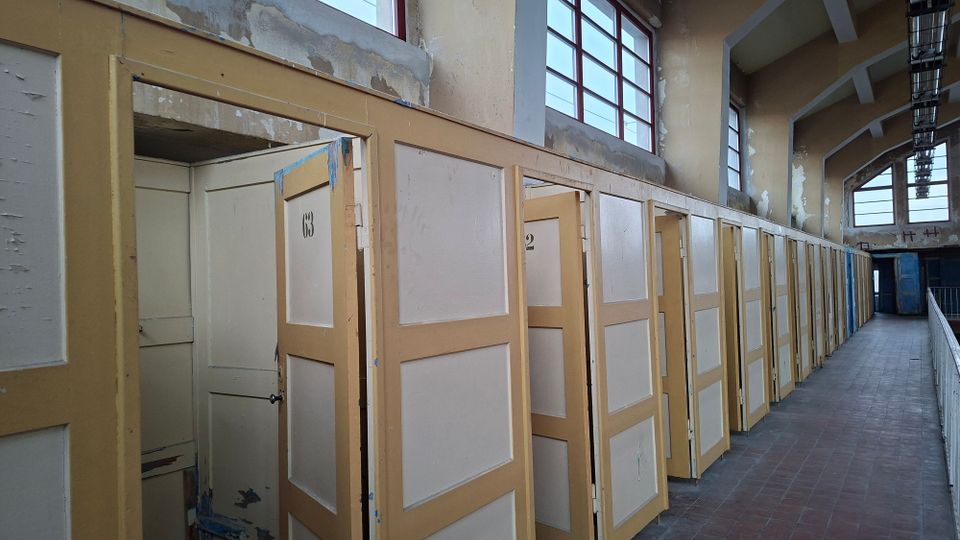 Ein alter Innenraum mit einer Reihe gelblicher Holzkabinen mit nummerierten Türen, darunter teilweise offene Türen. Die Wände sind verputzt und stellenweise abgenutzt, und oben verläuft eine Reihe von Fenstern mit roten Rahmen.