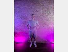 Indoor studio space with an exposed brick wall backdrop, lit by purple and pink uplights near the floor. Dark wood flooring and light haze create a soft, smoky effect across the lower part of the scene.