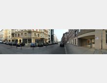 Straßenszene in einer städtischen Umgebung mit mehrstöckigen historischen und modernen Gebäuden auf beiden Seiten. Autos parken entlang der Straße, und Fußwege aus Pflastersteinen grenzen den Fahrbereich ab.