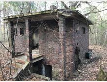 Verlassenes Backsteingebäude in einem bewaldeten Gebiet, mit verwitterten Wänden, zerbrochenen Fenstern und einer beschädigten Außentreppe. Das Gebäude ist von kahlen Ästen und fallendem Laub umgeben.