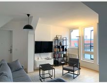 Modernes Wohnzimmer mit hellen Wänden und Holzboden, ausgestattet mit einem grauen Sofa, einem schwarzen Sessel und einem flachen Fernseher an der Wand. Große Fenster lassen viel Tageslicht herein, während ein Regal mit Büchern und Dekorationen sowie ein hängender schwarzer Lampenschirm den Raum ergänzen.