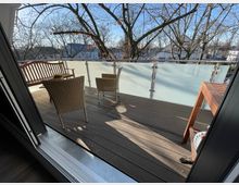 Eine Terrasse mit Holzboden und halbhohem, mattem Glasgeländer. Darauf stehen zwei Rattanstühle, eine Holzbank, ein kleiner Tisch mit Pflanze und ein weiterer Holztisch; im Hintergrund sind kahle Bäume und Wohnhäuser sichtbar.