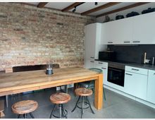 Moderne Küche mit rustikalem Stil: Sichtbare Ziegelwand, weißer Einbauschrank mit schwarzen Griffen, dunkle Arbeitsfläche, Herd und Mikrowelle. In der Mitte steht ein massiver Holztisch mit Holzbarstühlen auf einem grauen Fliesenboden.