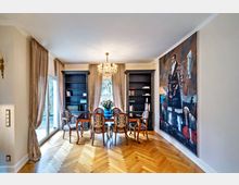 Ein elegantes Wohnzimmer mit einem Holzparkettboden und einem rechteckigen Holztisch, umgeben von fünf gepolsterten Stühlen. Große Fenster mit beigefarbenen Vorhängen lassen viel Tageslicht herein, flankiert von zwei schwarzen Bücherregalen. Ein Kronleuchter hängt von der Decke, und eine großformatige moderne Malerei bedeckt die rechte Wand.