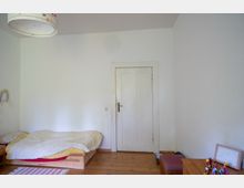 Schlichtes Schlafzimmer mit weiß gestrichenen Wänden, Holzdielenboden und einer zentralen weißen Tür. Links steht ein niedriges Bett auf Holzgestell mit Bettwäsche, rechts ein kleiner Tisch mit Gegenständen sowie eine Deckenlampe oben im Bild.