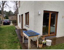 Einfamilienhaus mit hell verputzter Fassade, Holztüren und Fenstern mit Glaseinsätzen. Im Garten befinden sich ein Holzsteg, ein alter Gartentisch, mehrere Pflanztöpfe sowie eine Rasenfläche, daneben ein schwarzes geparktes Auto auf einer gepflasterten Einfahrt.