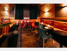 Gemütlicher Lounge- und Barbereich mit freigelegten Ziegelwänden, gepolsterten roten Sitzbänken entlang der Wände und kleinen runden Tischen mit Tischlampen. Samtstühle in Rot und Dunkelgrün stehen auf dunklem Boden, im Hintergrund hängen schwarze Vorhänge und an den Wänden sind Leuchten und Lautsprecher montiert.