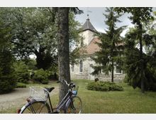 Eine kleine historische Kirche mit gemauerten Wänden und einem roten Ziegeldach, umgeben von hohen Bäumen und gepflegtem Grün. Im Vordergrund steht ein lila Fahrrad vor einem Baum auf einer Wiese.