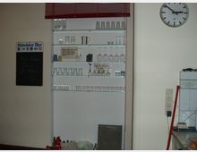 Ein schlicht eingerichteter Raum mit einem Regal, das verschiedene Gläser und Tassen ausstellt. An der Wand gibt es eine Uhr, eine Kreidetafel, und einen Lichtschalter, daneben sind weitere Haushaltsgegenstände sichtbar.