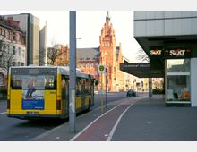 Straßenszene in einer städtischen Umgebung mit einem gelb-weißen Bus an einer Haltestelle vor einem Gebäude mit der Aufschrift „Bezirksamt Steglitz“. Im Hintergrund ein großes rotes Backsteingebäude mit Turm und historischen Details, umgeben von moderner und älterer Architektur.