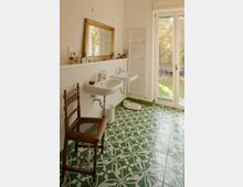 Helles Badezimmer mit zwei weißen Waschbecken, einem großen Spiegel mit goldfarbenem Rahmen und einer weißen Handtuchheizung an der Wand. Der Boden ist mit grünen und weißen Fliesen im dekorativen Muster gestaltet, und eine große Glastür führt zu einem Gartenbereich.