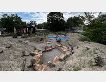 Ein offenes Gelände mit angelegten Beeten, Kieswegen und einer niedrigen Backsteinmauer. Im Hintergrund stehen Bäume, eine kleine schwarze Hütte und eine Gebäudegruppe mit Graffiti.