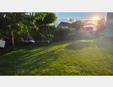 Garten mit großer Rasenfläche, umgeben von Hecken und Bäumen, mit mehreren Liegestühlen und einer Sitzbank. Tief stehende Sonne am Himmel wirft lange Schatten über den Rasen, im Hintergrund sind Hausdächer zu sehen.