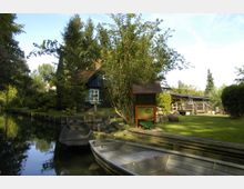 Ein idyllisches Ufer mit einem kleinen, dunkelblauen Holzhaus im Hintergrund, umgeben von Bäumen und einem Garten. Im Vordergrund liegen zwei Boote an einem schmalen Kanal, daneben eine Informationstafel und gepflegte Rasenflächen.