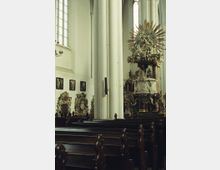 Innenraum einer historischen Kirche mit hohen, weißen Säulen, hölzernen Kirchenbänken und verzierten Statuen. Im Hintergrund steht ein reich dekorierter Altar mit goldenen Elementen und figürlicher Darstellung, flankiert von gotischen Fenstern mit farblosem Glas.