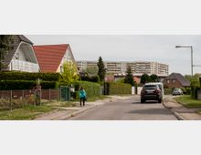Wohnsiedlung mit einer schmalen Straße, gesäumt von Einfamilienhäusern mit roten und dunklen Dächern sowie gepflegten Grünflächen. Im Hintergrund erheben sich mehrere mehrstöckige Plattenbauten, während eine Frau auf dem Gehweg entlanggeht und Autos am Straßenrand parken.