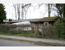 Ein kleiner, roter Schuppen mit Flachdach steht hinter einem Maschendrahtzaun und Büschen, daneben liegen Boote und Gartenmaterial. Im Vordergrund verläuft ein Gehweg, und ein Schild weist auf freie Liegeplätze hin.
