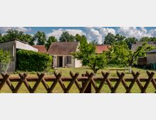 Ein kleiner Garten mit Rasenfläche und Obstbäumen, umgeben von einem Holzzaun. Im Hintergrund steht ein Einfamilienhaus mit roten Dachziegeln und cremefarbener Fassade, flankiert von weiteren Gebäuden und üppigem Grün.