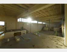 Verlassene Industriehalle mit rechteckigen Fenstern entlang der oberen Wände und sichtbaren Stahlträgern an der Decke. Im Vordergrund eine erhöhte Plattform mit Geländern und Treppen, die in den unteren Bereich der Halle führen; der Raum ist durch Sonnenlicht teilweise beleuchtet.