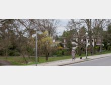 Straßenecke mit Straßenschildern „Gelfertstraße“ und „Auf dem Graf“, umgeben von kahlen Bäumen und einer grünen Wiese; im Hintergrund Wohnhäuser mit Balkonen. Eine Person spaziert mit einem kleinen Hund auf dem Bürgersteig.