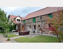 Hof mit einem renovierten, aus Stein und Ziegeln gebauten Gebäude mit roten Dachziegeln und grünen Holztüren, umgeben von einem Garten mit Bäumen und einem Gehweg aus Pflastersteinen. Im Vordergrund steht ein Whirlpool auf einer gepflasterten Fläche.