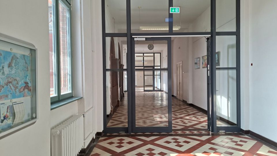 Langer Innenflur eines öffentlichen Gebäudes mit weiß gestrichenen Wänden, großen Fenstern links und einem gemusterten Fliesenboden in Rot- und Beigetönen. Im Vordergrund stehen schwarze Glasrahmentüren mit Fluchtweg-Schild, entlang des Flurs sind Heizkörper, Türen und gerahmte Bilder an der Wand zu sehen.