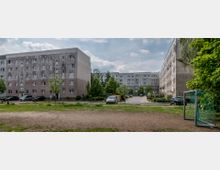 Wohnsiedlung mit mehreren grauen Plattenbauten in rechteckiger Anordnung, umgeben von Bäumen und Sträuchern. Im Vordergrund ein sandiger Bolzplatz mit einem Fußballtor, daneben geparkte Autos und ein Gehweg.