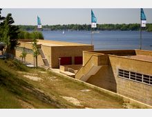 Modernes Gebäude mit beige-gelben Klinkerwänden und Treppenanlagen, gelegen an einem Hang mit Blick auf einen See. Im Hintergrund sind Wasser, Segelboote und die bewaldete Uferlinie erkennbar, während Fahnen auf dem Dach wehen.