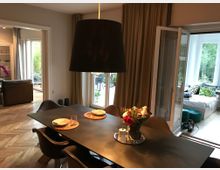 Essbereich in einem modernen Wohnraum mit einem rechteckigen Tisch, auf dem Teller und Blumen stehen. Im Hintergrund sind ein Wohnzimmer mit Sofa sowie ein Wintergarten mit Glasfenstern und Blick ins Grüne erkennbar.