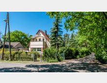 Zweistöckiges Einfamilienhaus mit rotem Satteldach und kleinem Balkon, umgeben von einem gepflegten Garten und einem Zaun. Rechts führt ein schattiger Weg durch eine Baumallee, während das Haus von Bäumen und dichter Vegetation umgeben ist.