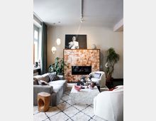 Wohnzimmer mit gemütlicher Einrichtung: Zentral steht ein Kamin mit marmorierter Verkleidung und einem Schwarz-Weiß-Porträt darüber. Umgeben ist der Raum von einem grauen Sofa, weißen Sesseln, einem Couchtisch aus hellem Marmor sowie Pflanzen und dekorativen Lampen. Ein Teppich mit feinem Rautenmuster bedeckt den Boden.