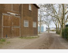 Holzgebäude mit einer großen Holztür und mehreren Fenstern auf der linken Seite, daneben ein unbefestigter Weg, der von kahlen Bäumen gesäumt ist. Im Hintergrund ist ein weiß-blaues Gebäude sowie ein Geländer zu sehen.