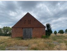 Backstein-Scheune mit steilem Satteldach und einer Holztür auf der Vorderseite, umgeben von einer Grasfläche und vereinzelt stehenden Bäumen, unter einem bewölkten Himmel. Auf der Fassade sind die Buchstaben "F M" in das Mauerwerk integriert.