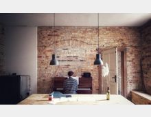 Ein Raum mit sichtbarer Backsteinwand und einer alten Holztür im Hintergrund. Zentral spielt eine Person Klavier, davor steht ein Holztisch mit Papieren und einer Vase, beleuchtet von zwei hängenden schwarzen Lampen.