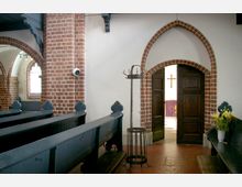 Innenraum einer Kirche mit roten Ziegelbögen und hölzernen Bänken. Ein geöffneter Rundbogen aus Ziegeln führt zu einem Nebenraum, in dem ein Holzkreuz und ein lila Altartuch sichtbar sind; rechts steht eine Bank mit einem Blumenstrauß in einer Vase.